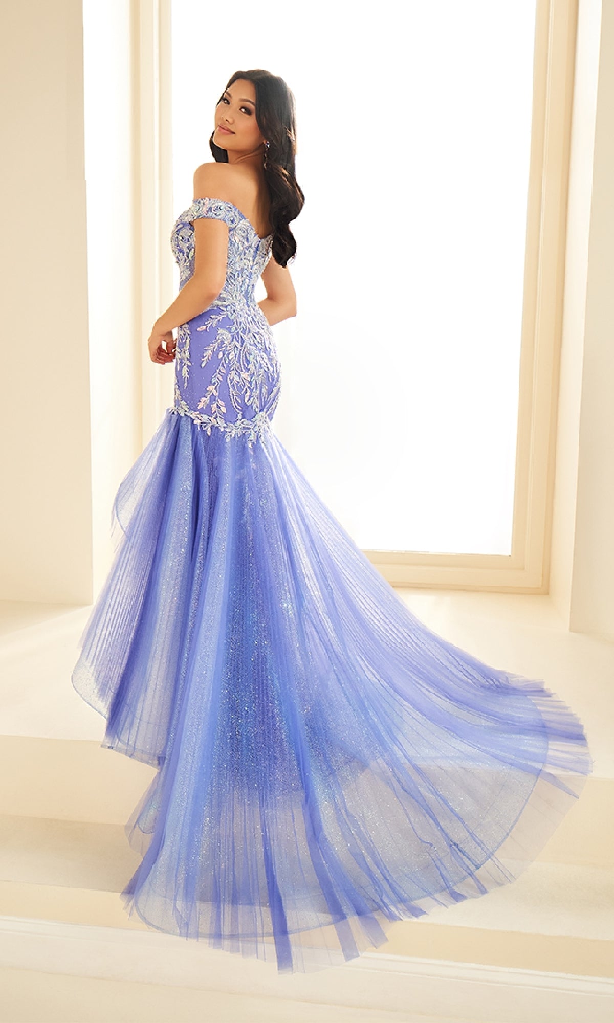 A woman stands on steps wearing the Ellie Wilde EW36036, a fitted lavender off-the-shoulder prom dress with silver embroidery and a tulle mermaid skirt, looking over her shoulder.